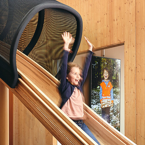 Ein Mädchen rutscht auf einer hölzernen Rutsche im NAWAREUM. Sie lacht und hat die Hände hochgehoben. Der Innenraum ist ganz mit Holz verkleidet.