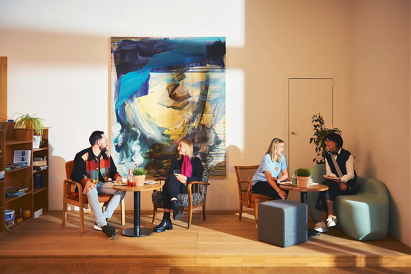 Vier Menschen sitzen jeweils zu zweit an Café-Tischen und unterhalten sich. Sie haben Getränke und kleine Speisen vor sich. Der Raum wird von warmem Licht von draußen erhellt. Im Hintergrund hängt ein abstraktes Gemälde mit viel Blau.