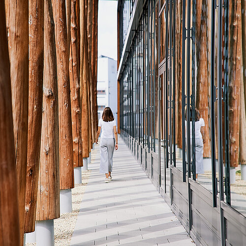 Links eine Reihe rotbrauner Baumstämme, die das Vordach des NAWAREUMs tragen. Rechts die Fensterfront des NAWAREUMs. In der Mitte geht eine weiß gekleidete Frau.