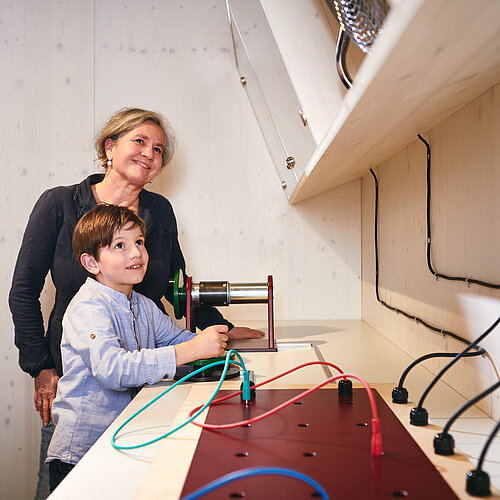 Ein Junge kurbelt an einer Mitmachstation im NAWAREUM um Strom zu erzeugen. Seine Oma schaut ihm neugierig zu.