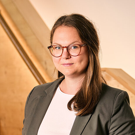 Porträt einer braunhaarigen Frau mit Brille. Sie steht vor dem Handlauf einer Holztreppe.