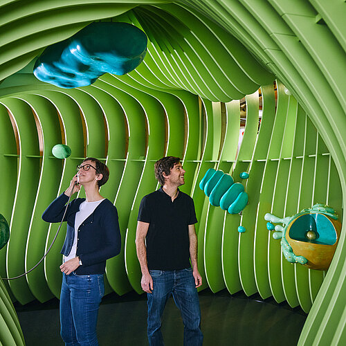 Ein Mann und eine Frau stehen und staunen in dem überdimensionalen Modell einer Pflanzenzelle. Die Frau hält einen Einhandhörer an ihr Ohr. Die Zellwände und die Zelllorganellen sind grün dargestellt.