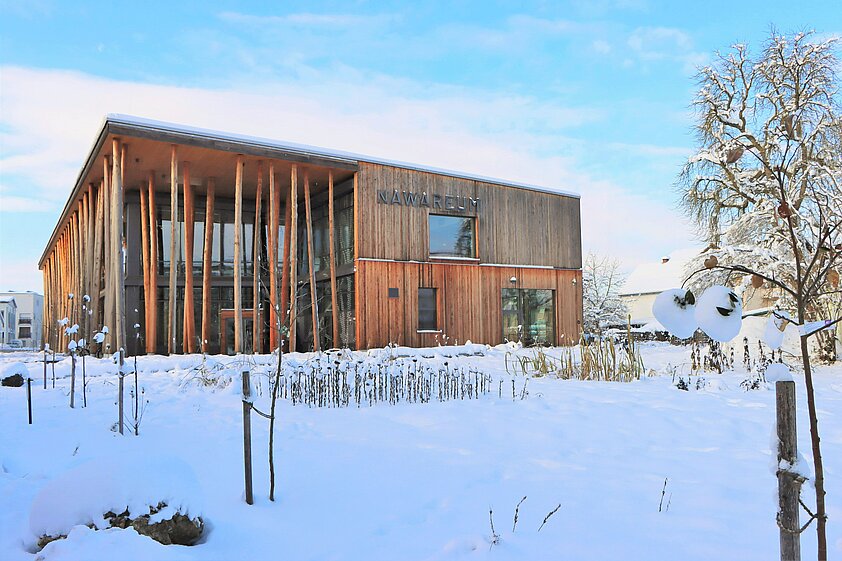 Das NAWAREUM - ein großes, quaderförmiges Holzgebäude - steht inmitten eines schneebedeckten Gartens.