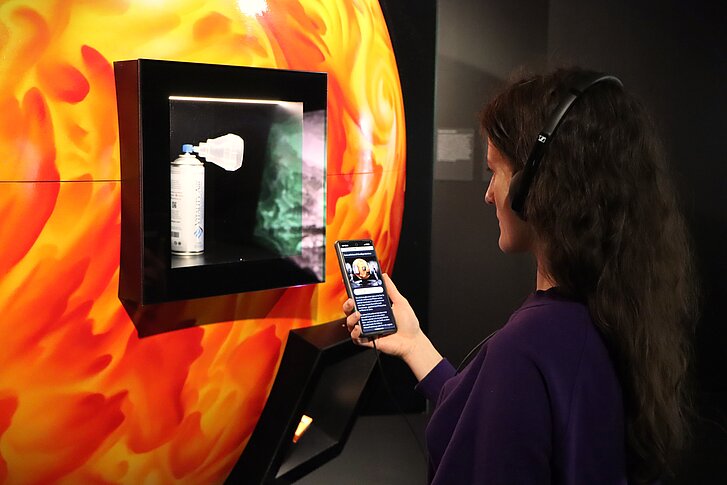 Eine junge Frau mit Kopfhörern steht vor einem Modell der Sonne und blickt auf das Handy in ihrer Hand.