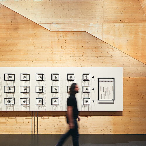 Im NAWAREUM geht ein Mann durch das Bild. Er befindet sich in einem hohen Raum mit Holzverkleidung. An der Wand hängt eine künstlerische Installation mit dunklen Würfelgerüsten auf weißem Hintergrund.